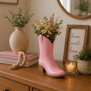 May include: A pink cowboy boot vase filled with dried flowers sits on a wooden dresser. A beige vase with greenery, a framed sign that says "home sweet home", and a lit candle are also on the dresser. Two pink books and a string of beads are also visible.