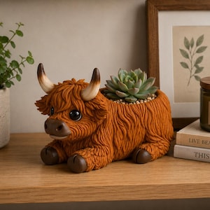 May include: A brown Highland cow planter with a succulent plant. The ceramic planter has a shaggy texture and features a detailed face with large eyes and horns. The planter is sitting on a wooden surface.