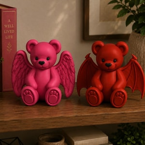 May include: Two sitting bear figurines, one bright pink with feathered angel wings and one deep red with pointed bat wings. Both bears have black eyes and noses, rounded ears, and paw-pad details on their feet, displayed side by side.