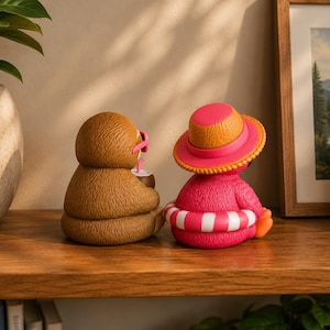 May include: Two sloth figurines on a wooden shelf. One is brown, holding a drink with pink flamingo sunglasses. The other is pink, wearing a hat and a white and pink striped floatie. A framed landscape painting is in the background.