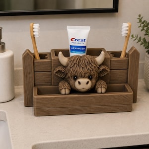 May include: A brown bathroom organiser shaped like a wooden fence with a sculpted Highland cow figure in the centre. It features two side compartments for toothbrushes and a central slot holding a tube of Crest Pro-Health Advanced toothpaste.