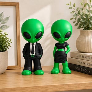 May include: Two green alien figurines with large black eyes. One figure wears a black suit with a white shirt and black tie. The other figure wears a black crop top and a black skirt. Both have green skin and stand on a flat surface.