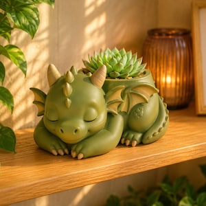 Può includere: Un vaso a forma di drago verde con una pianta succulenta. Il drago è in posizione di riposo, con gli occhi chiusi, piccole corna e ali. Il vaso è su una mensola di legno, con una candela e un'opera d'arte incorniciata sullo sfondo.