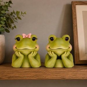Peut inclure: Deux figurines de grenouilles vertes aux grands yeux sombres et aux joues roses, toutes deux posant leur menton sur leurs mains. La grenouille de gauche porte un petit nœud rose sur la tête, tandis que celle de droite porte un petit nœud papillon noir.