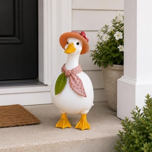 May include: A whimsical white goose statue with a yellow beak and feet. It wears an orange hat with a red bow, a pink scarf with white polka dots, and a green leaf. The goose is standing on a concrete step.