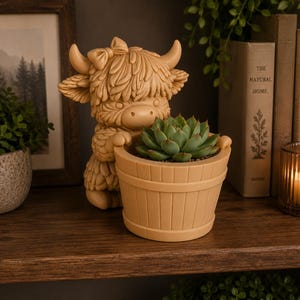 May include: A light tan resin planter shaped like a shaggy Highland cow with a bow on its head, holding a round, barrel-style pot. A green succulent plant is potted inside the container, which features vertical slats and a smooth, matte finish.