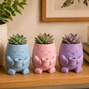 May include: Three small, rounded planters in light blue, soft pink, and lavender, each featuring a sculpted character face with arms holding a small cup. Each planter contains a green or purple succulent plant, creating a set of three distinct, colorful, and cheerful plant pots.