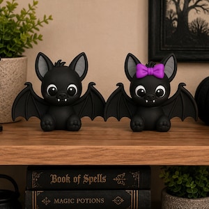 May include: Two small black bat figurines with large white eyes and visible fangs. One bat features a bright purple bow on its head. Both figures have outstretched wings, rounded ears with grey inner details, and are positioned side by side.