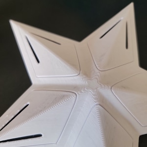 K&ouml;nnte beinhalten: Ein wei&szlig;es, sternf&ouml;rmiges Objekt mit f&uuml;nf Spitzen und einer strukturierten Oberfl&auml;che. Das Design weist kleine, rechteckige Aussparungen an den Kanten jeder Spitze auf. Es scheint ein dekoratives Element oder Teil einer gr&ouml;&szlig;eren Struktur zu sein.