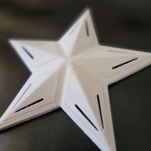 K&ouml;nnte beinhalten: Ein wei&szlig;es, sternf&ouml;rmiges Objekt mit f&uuml;nf Zacken und einem geometrischen Design. Der Stern hat eine strukturierte Oberfl&auml;che und mehrere rechteckige Aussparungen entlang der Kanten jeder Spitze. Das Objekt scheint aus einem festen Material zu bestehen.