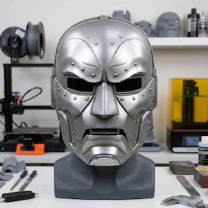 May include: A metallic silver full-face mask featuring a segmented design with visible rivets along the brow, cheeks, and jawline. The mask has rectangular eye openings and a horizontal mouth slit, displayed on a dark gray mannequin head bust.