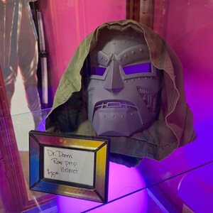 May include: A grey Dr. Doom raw prop helmet with a green hood. The helmet has purple eye lenses and is displayed with a framed sign that reads "Dr. Doom Raw prop Helmet".
