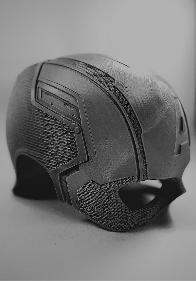 Pu&ograve; includere: Un casco grigio scuro con pannelli strutturati e un design dettagliato. Il casco presenta una combinazione di superfici lisce e decorate, con accenti angolari e una forma curva. L'estetica generale suggerisce un tema futuristico o tattico.