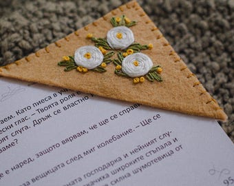 May include: A tan felt bookmark with three embroidered white flowers with yellow centers and green leaves. The bookmark is triangular and rests on a book page.