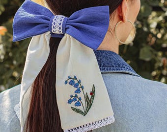May include: A blue linen hair bow with a white lace center and long, cream-colored tails. The tails are embroidered with blue flowers and a ladybug, trimmed with lace. The bow is worn in a ponytail.