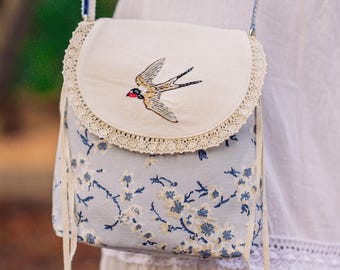 May include: A small white and blue floral patterned crossbody bag with a white lace trim and a small embroidered bird on the front flap. The bag has a long strap and a drawstring closure.