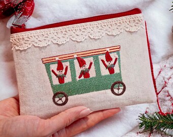 May include: A beige fabric pouch with a red zipper and trim, adorned with a cream lace border. The pouch features an embroidered green train car with three elves wearing red hats. The pouch is held against a white, textured background.
