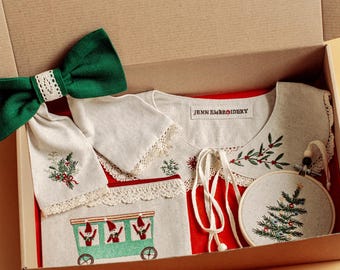 May include: A Christmas-themed gift set in a cardboard box. The set includes a white and red embroidered top with a train design, a green bow with lace trim, and a small embroidered Christmas tree ornament. The text "JENN EMBROIDERY" is visible.