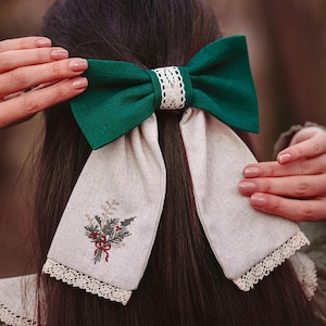 May include: A dark green bow hair accessory with a white lace centre and long, cream-coloured tails. The tails are embroidered with a floral design and trimmed with lace. The accessory is worn in the hair.