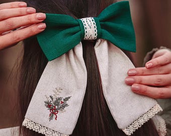 May include: A dark green bow hair accessory with a white lace center and long, cream-colored tails. The tails are embroidered with a floral design and trimmed with lace. The accessory is worn in the hair.