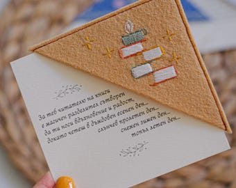May include: A handmade bookmark with a tan felt triangle featuring embroidered books and a candle. The bookmark is held over a card with text in a script font. The card has a floral design.