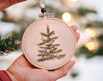 May include: Embroidered Christmas tree ornament in a wooden hoop. The tree is green with red, blue, and yellow ornaments. The ornament is held by a person's hands, with a cream-colored string for hanging.