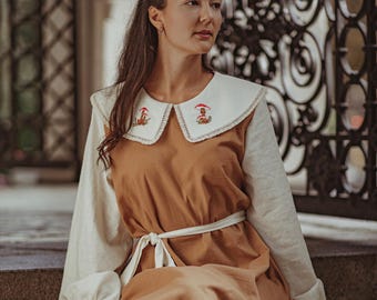 May include: A long, brown dress with a white collar and sleeves. The collar features embroidered mushroom designs. The dress has a white tie belt and a ruffled hem. The sleeves have ruffled cuffs.