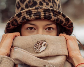 May include: A person wearing a leopard print bucket hat and a tan scarf with a hedgehog brooch. The brooch is a detailed embroidery with brown and white thread. The background is blurred with neutral tones.