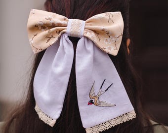 May include: A large hair bow with a floral pattern in beige and white, paired with lavender fabric tails. The tails are adorned with a delicate embroidered swallow and lace trim, creating a charming accessory.