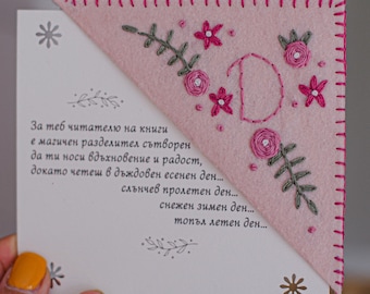 May include: Pink felt bookmark with embroidered floral design and the letter "D". The bookmark has a stitched border and is placed on a white card with text in a foreign language. The bookmark is held by a hand.