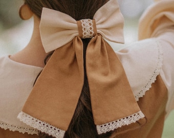 May include: A large, two-toned hair bow in cream and tan, with a delicate lace accent at the center and along the edges of the long, flowing tails. The bow is tied in the hair, adding a touch of elegance.