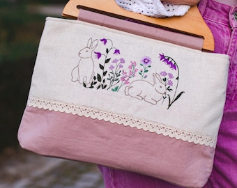 May include: A cream-colored tote bag with a wooden handle. The bag features an embroidered design of two rabbits and purple flowers. The bottom half of the bag is a dusty rose color, with a cream lace trim.