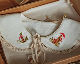 May include: Cream-colored Peter Pan collar with embroidered mushroom designs. The collar features a delicate lace trim along the edge and a tie closure. The collar is presented in a wooden box.
