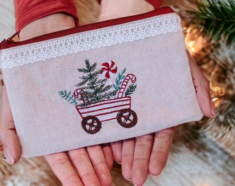 May include: A beige rectangular pouch with a red zipper and a white lace trim. The pouch features an embroidered Christmas design of a wagon carrying a Christmas tree, candy canes, and other festive elements.