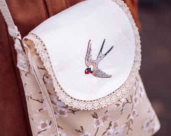 May include: A beige fabric crossbody bag with a white flap featuring an embroidered swallow. The bag has a floral print and a brown tassel.