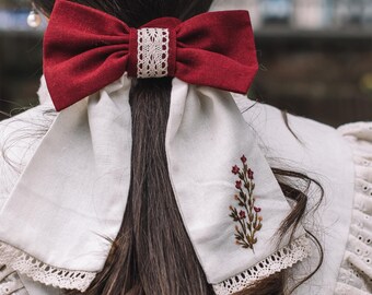 May include: A burgundy bow hair accessory with a white lace center is secured in dark brown hair. The bow sits atop a white fabric piece with a floral embroidery and lace trim, creating a vintage aesthetic.