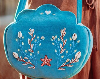 May include: A blue velvet handbag shaped like a seashell with embroidered white seashells, coral, and a red starfish.