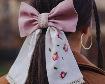 May include: A large, dusty rose bow with a lace center is attached to a white fabric tail. The tail is embroidered with pink and yellow flowers and green leaves. The bow is a hair accessory.