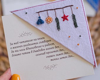 May include: A handmade white felt bookmark with embroidered holiday-themed decorations, including a blue tassel, orange slice, red star, and green sprigs. The bookmark is placed on a cream-colored card with text.