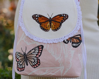 May include: A small, pink crossbody bag with a butterfly design. The bag features embroidered butterflies in shades of orange, brown, and pink. The flap is cream-colored with white lace trim. The bag has a white drawstring closure.