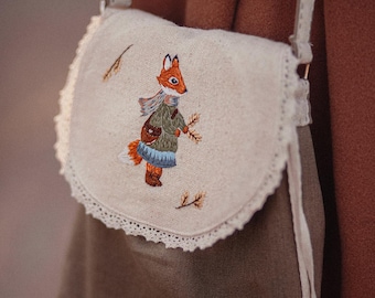 May include: A small, light brown canvas bag with a white lace trim. The bag has a fox embroidered on the front, wearing a blue dress and holding a branch. The bag has a long strap.