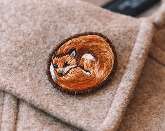 May include: A brown embroidered patch of a curled up orange fox with white accents. The patch is sewn onto a brown fabric.
