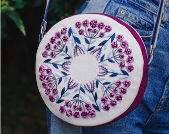 May include: A round white felt bag with a purple trim and a floral embroidery design in shades of purple and blue. The bag has a long strap.