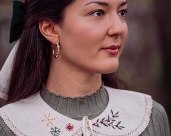 May include: A woman wearing a green sweater and a cream-colored embroidered collar. The collar features a snowflake, a Christmas tree, and a red berry design. The collar is trimmed with lace and has a tie closure.