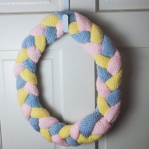 May include: A braided wreath in pastel colors of yellow, pink, and blue. The wreath is made of a soft, knitted material and is hanging on a white door. The wreath is a decorative item.
