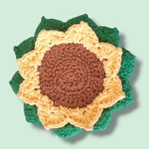 May include: A crocheted sunflower coaster with green leaves and a brown centre. The coaster is made of yarn and has a textured surface.