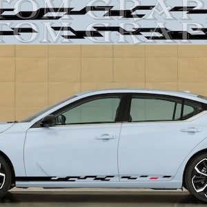 Peut inclure: Une berline bleu clair avec des bandes de course noires et rouges le long des côtés. La voiture a des roues noires et une calandre noire. La voiture est garée devant un mur beige. "CUSTOM GRAPHICS" est écrit en lettres noires et rouges sur le mur.