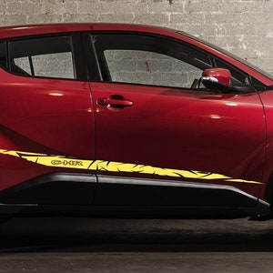 Op de afbeelding: Een rode Toyota C-HR met zwarte wielen en gele en zwarte race strepen langs de zijkant. De auto staat geparkeerd op een grijs betonnen oppervlak.