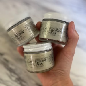 May include: Three small glass jars of deodorant cream are held in a hand. Each jar has a white lid and a label with the product name and scent: Rosemary Mint, Orange Patchouli, and Blue Tansy Rose. The jars are 2.4 oz (68 g).