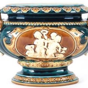 Op de afbeelding: Een sierlijke, teal-kleurige keramische vaas met gouden en bruine accenten. De vaas heeft een gedetailleerd reliëf van cherubijnen in een bruin ovaal kader. Decoratieve handvatten met leeuwenkoppen en ingewikkelde patronen sieren de zijkanten.